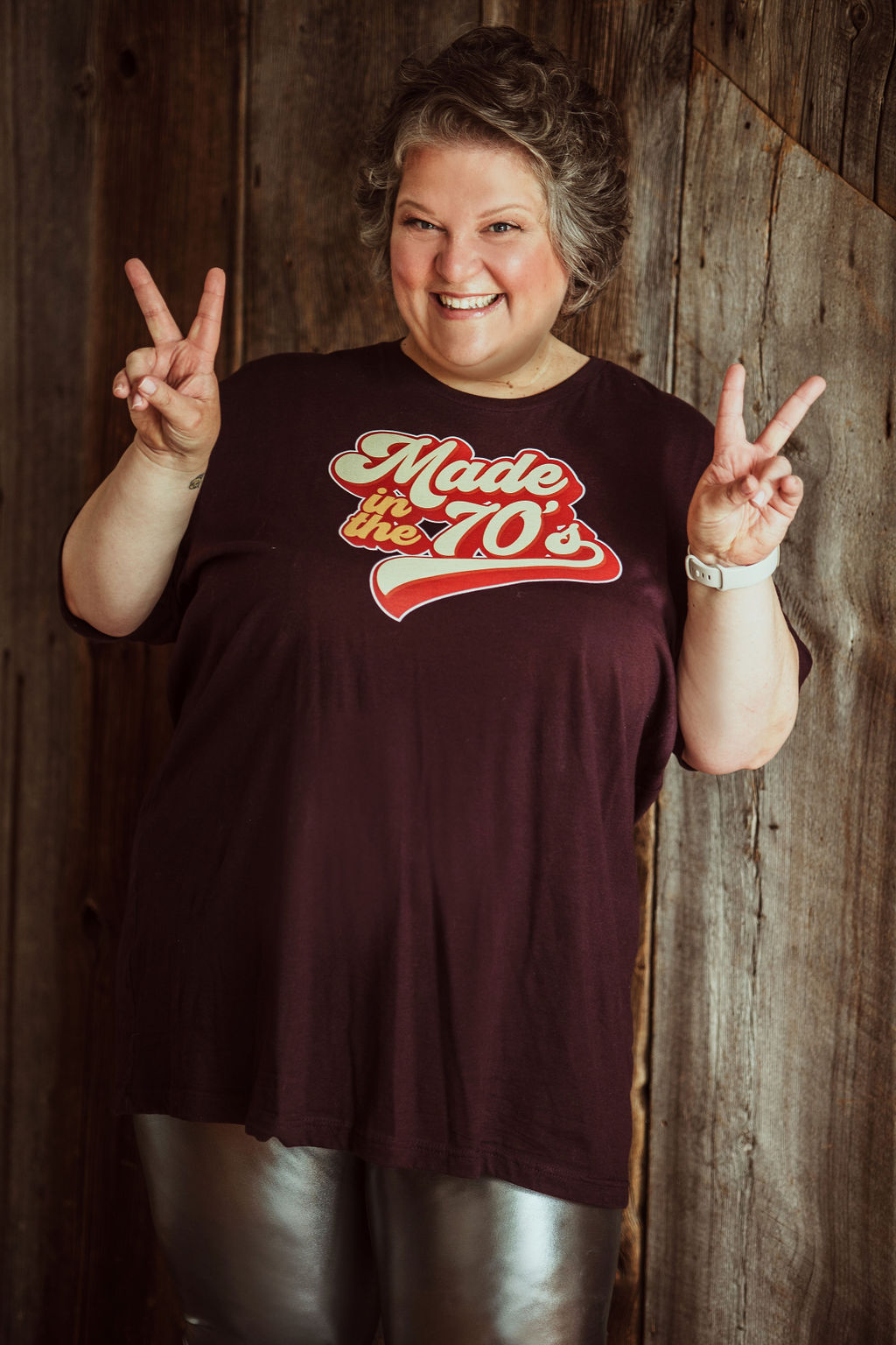 A woman giving the peace sign with "Made in the 70s" on her purple t shit, she is also wearing silver tights. She is celebrating her life.